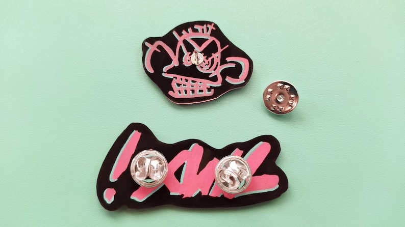 May include: Two enamel pins with black outlines and pink fill. One pin features a cartoon face with the text "!" and the other pin features the word "YAWL".