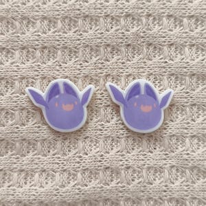 May include: Two lavender-colored, cartoon-style stickers with a smiling face and wing-like ears. The stickers are on a cream-colored, textured fabric background. The stickers have a white border.