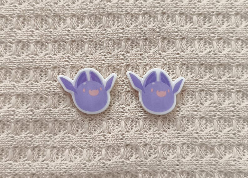May include: Two lavender-colored, cartoon-style stickers with a smiling face and wing-like ears. The stickers are on a cream-colored, textured fabric background. The stickers have a white border.