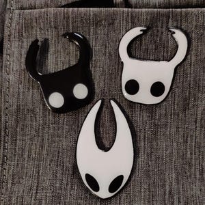 Może przedstawiać: Trzy emaliowane przypinki przedstawiające postacie z gry wideo Hollow Knight. Przypinki są czarne i białe i przedstawiają Rycerza, Straż Kr&oacute;lowej i Mistrza Duszy.