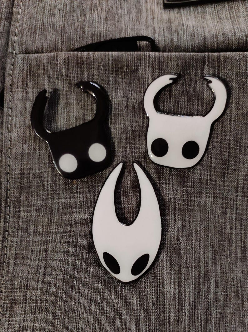 Może przedstawiać: Trzy emaliowane przypinki przedstawiające postacie z gry wideo Hollow Knight. Przypinki są czarne i białe i przedstawiają Rycerza, Straż Kr&oacute;lowej i Mistrza Duszy.