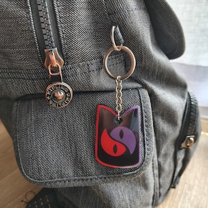 May include: A black, red, and purple cat-shaped keychain hanging from a silver keyring attached to a gray backpack. The keychain features a yin-yang design with cat eyes. The backpack has a zipper and a small pocket.