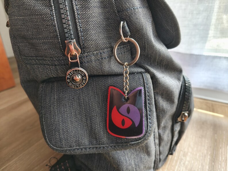 May include: A black, red, and purple cat-shaped keychain hanging from a silver keyring attached to a gray backpack. The keychain features a yin-yang design with cat eyes. The backpack has a zipper and a small pocket.