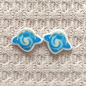 May include: Two blue and white enamel pins with a celestial design. Each pin features a blue planet shape with a white swirl and small white stars. The pins are set against a beige knitted background.