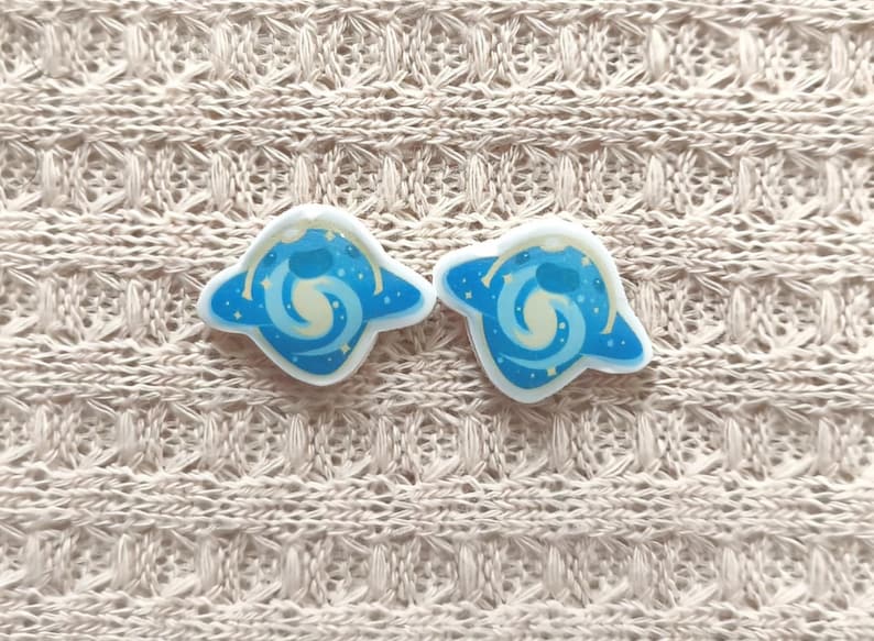 May include: Two blue and white enamel pins with a celestial design. Each pin features a blue planet shape with a white swirl and small white stars. The pins are set against a beige knitted background.