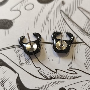 May include: A pair of black stud earrings with a curved, horn-like design. The earrings have a shiny, textured surface and a silver post and backing. The earrings are displayed on a white background with black line art.