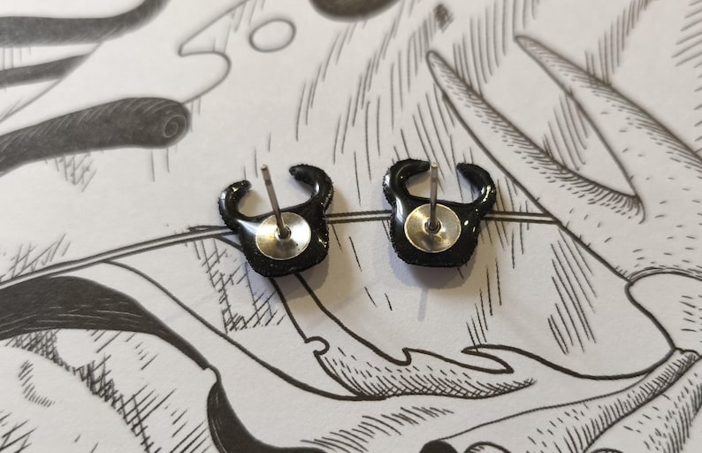 May include: A pair of black stud earrings with a curved, horn-like design. The earrings have a shiny, textured surface and a silver post and backing. The earrings are displayed on a white background with black line art.