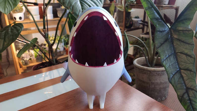 May include: A cartoon shark figurine with a wide open mouth, white teeth, and a purple interior. The shark has a white body, blue fins, and small legs. It is standing on a wooden surface, with plants in the background.
