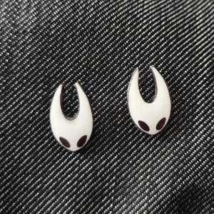 May include: Two white enamel pins with a stylized, curved shape and two small, dark purple dots. The pins are set against a textured, dark gray background, creating a striking contrast. The design is simple yet distinctive.