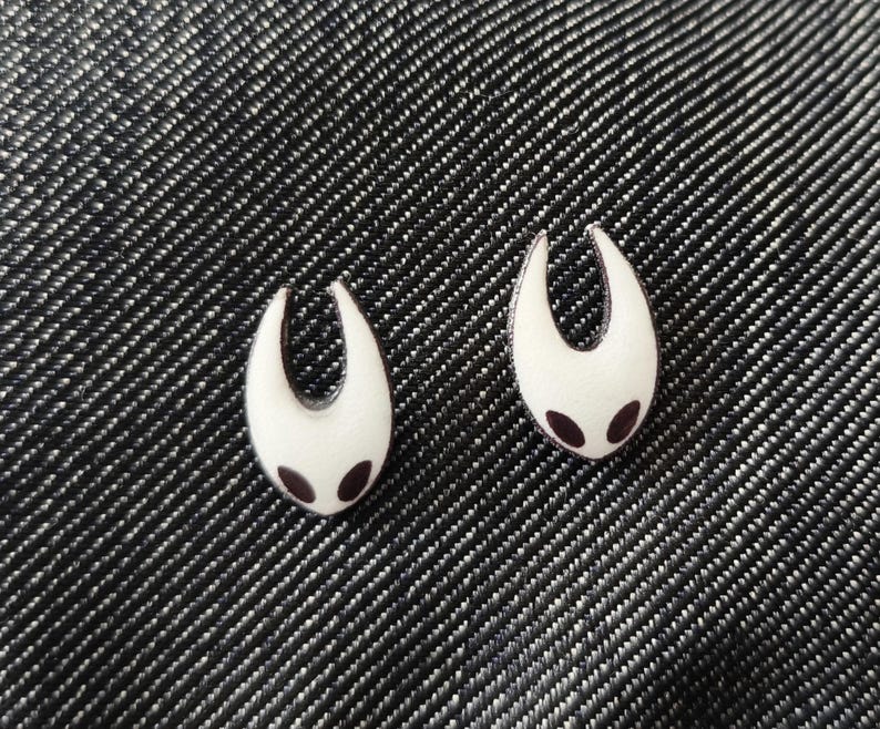 May include: Two white enamel pins with a stylized, curved shape and two small, dark purple dots. The pins are set against a textured, dark gray background, creating a striking contrast. The design is simple yet distinctive.
