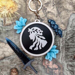 May include: A black and white keychain with a woman's silhouette, a blue sword, and blue floral accents. The keychain is round and has a silver keyring. Additional blue decorative elements and a small blue object with text are also present.