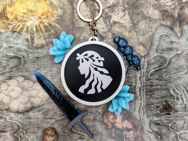 May include: A black and white keychain with a woman's silhouette, a blue sword, and blue floral accents. The keychain is round and has a silver keyring. Additional blue decorative elements and a small blue object with text are also present.