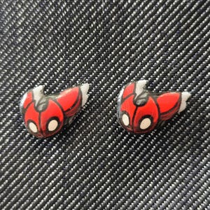 May include: Two small, red and black ladybug-shaped earrings with white accents. The earrings are set against a textured, dark gray background. These earrings are a fun accessory.