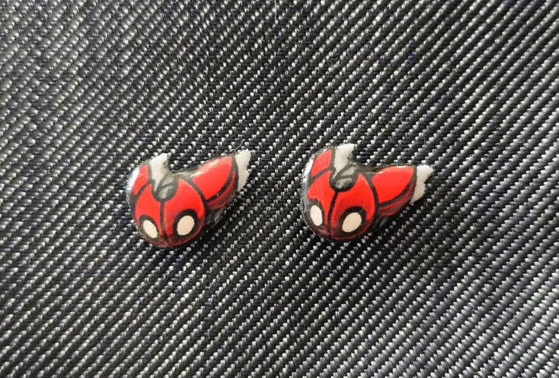 May include: Two small, red and black ladybug-shaped earrings with white accents. The earrings are set against a textured, dark gray background. These earrings are a fun accessory.