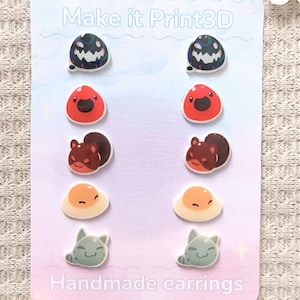 May include: A set of handmade earrings on a light pink card. The earrings feature various designs, including a dark green and black blob, a red blob, a brown squirrel, a fried egg, and a gray cat. The card reads "Make it Print3D" and "Handmade earrings."