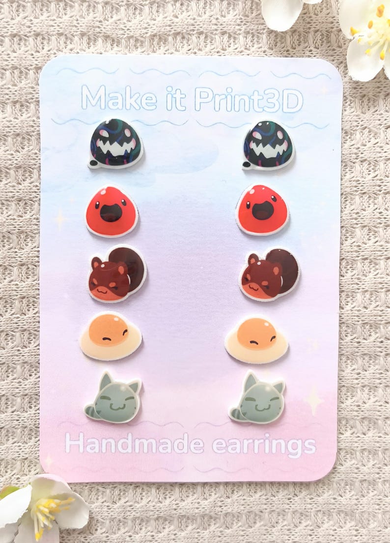 May include: A set of handmade earrings on a light pink card. The earrings feature various designs, including a dark green and black blob, a red blob, a brown squirrel, a fried egg, and a gray cat. The card reads "Make it Print3D" and "Handmade earrings."