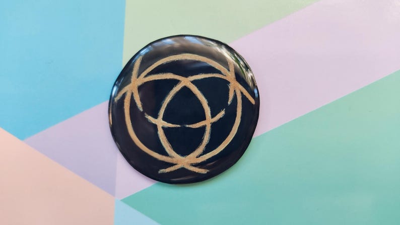 May include: A black circular coaster with a gold geometric design. The coaster has a smooth, glossy finish.