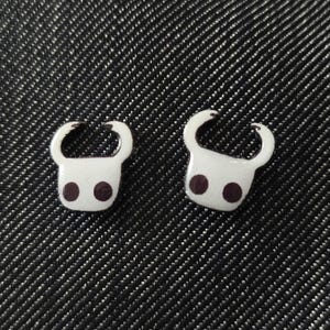 May include: Two white enamel pins, each featuring a stylized head with curved horns and two dark purple dots. The pins are set against a dark, textured background.
