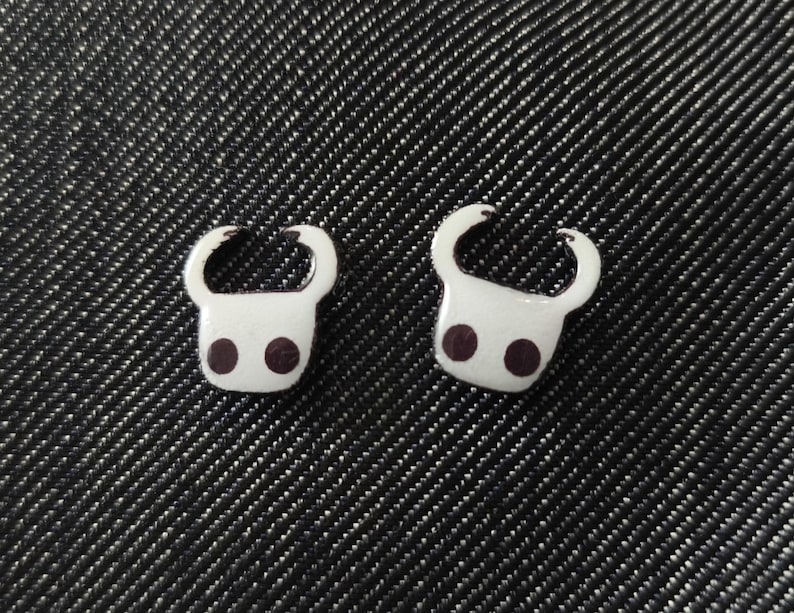 May include: Two white enamel pins, each featuring a stylized head with curved horns and two dark purple dots. The pins are set against a dark, textured background.