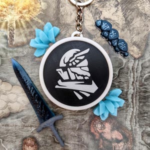 May include: A black and white round keychain with a stylized graphic, a blue sword, and blue flower-shaped decorations. The keychain has a silver chain and is set against a map-like background.