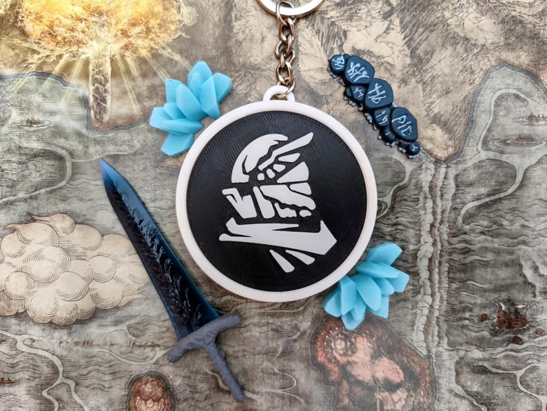 May include: A black and white round keychain with a stylized graphic, a blue sword, and blue flower-shaped decorations. The keychain has a silver chain and is set against a map-like background.