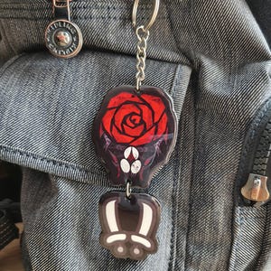 May include: A two-part keychain featuring a red rose design on a black background, and a cartoon bunny paw. The keychain is attached to a silver chain and a silver keyring. The keychain is hanging on a denim backpack.
