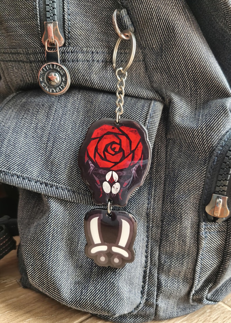 May include: A two-part keychain featuring a red rose design on a black background, and a cartoon bunny paw. The keychain is attached to a silver chain and a silver keyring. The keychain is hanging on a denim backpack.