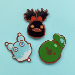 May include: Three enamel pins featuring cartoon characters. The first pin is black with a red bow and a toothy grin. The second pin is light blue with white accents and large eyes. The third pin is green with a brown face and a small mouth.