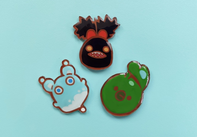 May include: Three enamel pins featuring cartoon characters. The first pin is black with a red bow and a toothy grin. The second pin is light blue with white accents and large eyes. The third pin is green with a brown face and a small mouth.