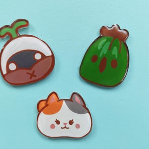 May include: Three colorful enamel pins featuring a green watermelon, a brown and white mushroom, and a white cat with orange and gray markings.