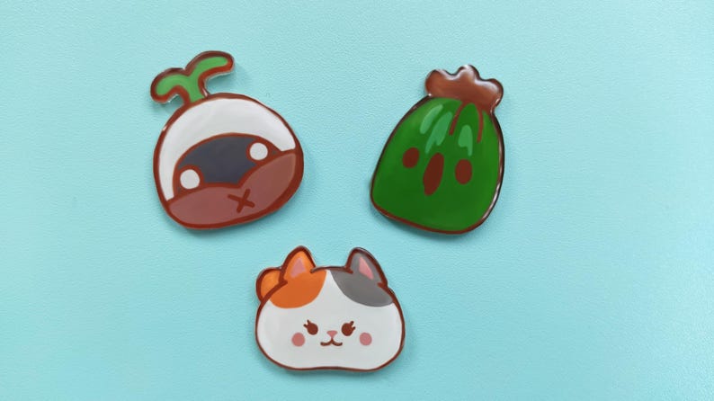 May include: Three colorful enamel pins featuring a green watermelon, a brown and white mushroom, and a white cat with orange and gray markings.