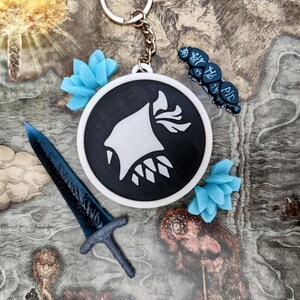 May include: A black and white keychain with a circular pendant featuring a stylized white design. Accompanying the keychain are a blue sword, blue floral accents, and a dark blue charm with text. The items are arranged on a map.
