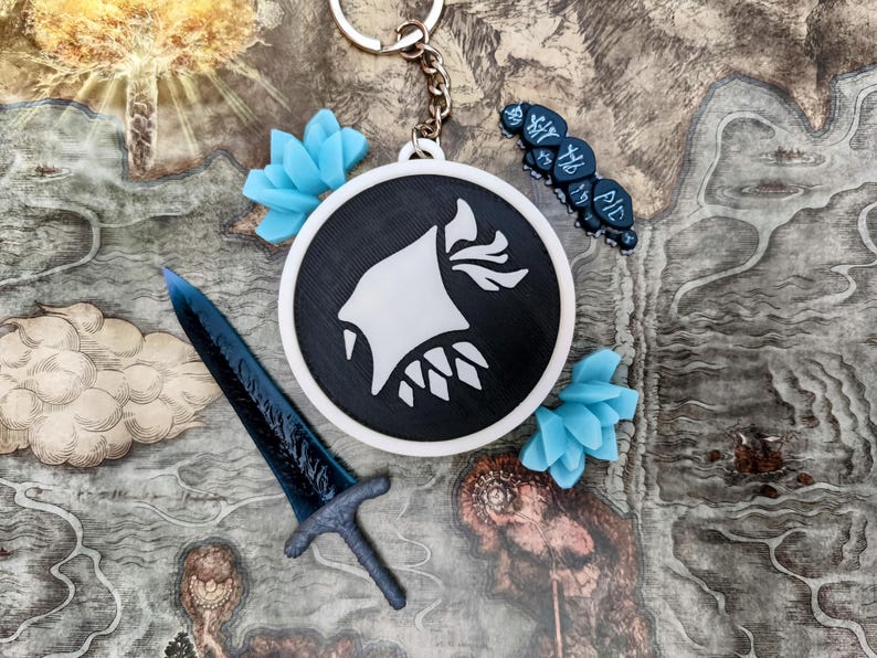May include: A black and white keychain with a circular pendant featuring a stylized white design. Accompanying the keychain are a blue sword, blue floral accents, and a dark blue charm with text. The items are arranged on a map.