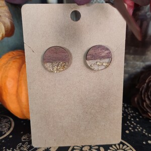 May include: A pair of round wooden earrings with a brown and white wood grain pattern and gold glitter embedded in clear resin.