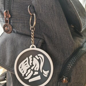 May include: A black and white round keychain with a stylized image of a woman's face and hair. The keychain is attached to a silver chain and a denim backpack with a zipper.