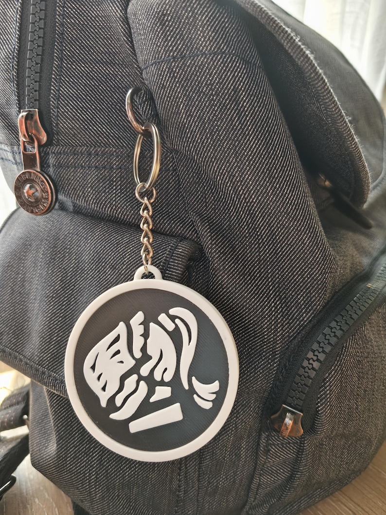 May include: A black and white round keychain with a stylized image of a woman's face and hair. The keychain is attached to a silver chain and a denim backpack with a zipper.