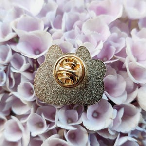 May include: Gold-colored enamel pin with a detailed design. The pin has a safety clasp on the back.