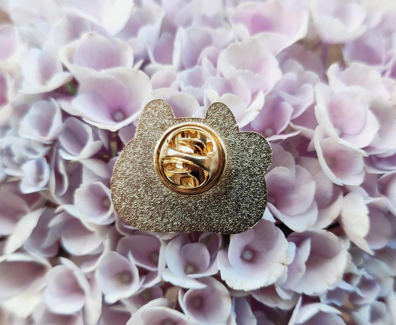 May include: Gold-colored enamel pin with a detailed design. The pin has a safety clasp on the back.