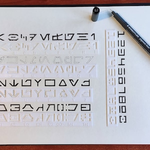 Peut inclure: Un carnet blanc avec une reliure spirale et un stylo noir pos&eacute; dessus. Le carnet a une grille de symboles en relief blancs sur le devant.