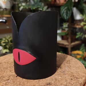 Op de afbeelding: Zwarte keramische plantenpot met een rood oogontwerp. De pot heeft de vorm van een cilinder met een licht uitlopende bovenkant.