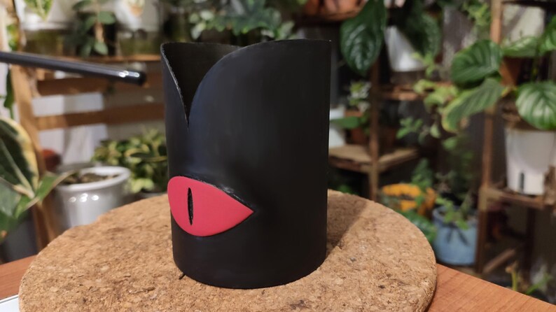 Op de afbeelding: Zwarte keramische plantenpot met een rood oogontwerp. De pot heeft de vorm van een cilinder met een licht uitlopende bovenkant.