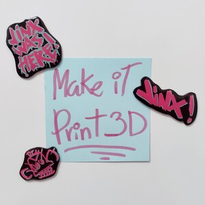 May include: A blue sticky note with the text "Make it Print 3D" written in pink. There are four pink and black stickers with various text on a white background.