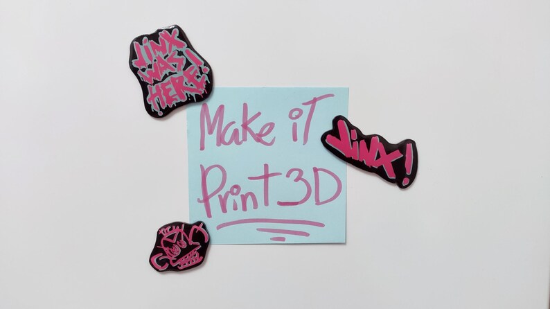 May include: A blue sticky note with the text "Make it Print 3D" written in pink. There are four pink and black stickers with various text on a white background.