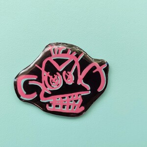 May include: A black and pink enamel pin with a cartoon skull design. The skull has a pink outline and white eyes.