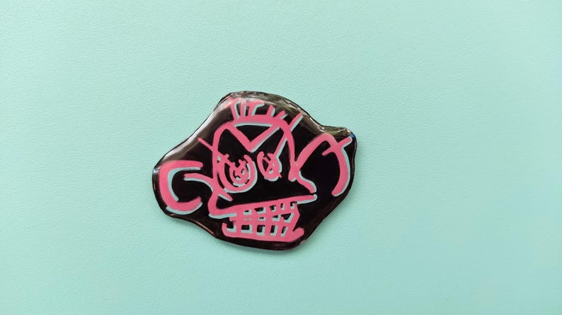 May include: A black and pink enamel pin with a cartoon skull design. The skull has a pink outline and white eyes.