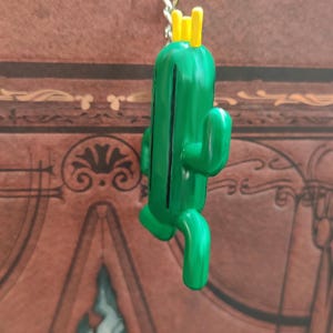May include: A green cactus keychain with a yellow crown on top. The cactus is standing upright with its arms outstretched.