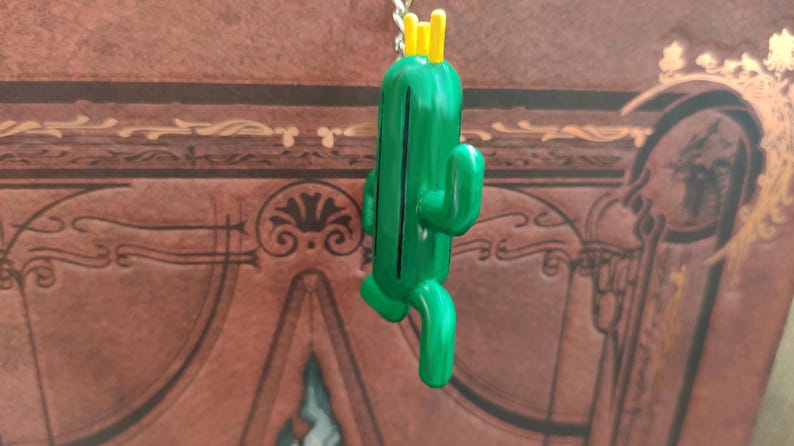 May include: A green cactus keychain with a yellow crown on top. The cactus is standing upright with its arms outstretched.