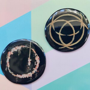 May include: Two round black and gold pinback buttons. One button features a gold abstract design on a black background. The other button features a white and pink abstract design on a black background.