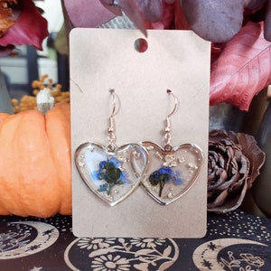 May include: A pair of heart-shaped earrings with blue flowers and gold flakes encased in clear resin. The earrings are on a brown card.