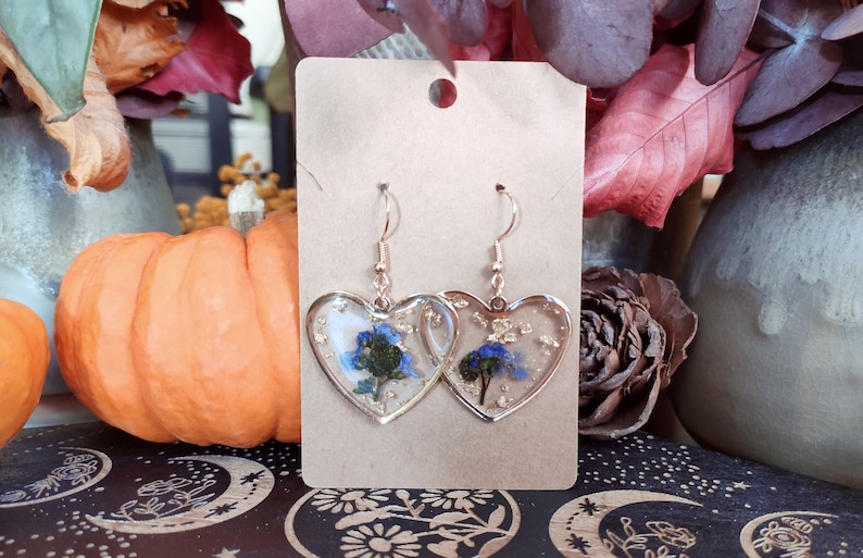 May include: A pair of heart-shaped earrings with blue flowers and gold flakes encased in clear resin. The earrings are on a brown card.